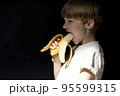 Funny portrait of boy with blond hair eating banana on black background. 95599315