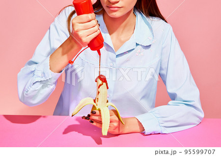 Food pop art photography. Young woman tasting banana with tomato ketchup isolated on pink background. Vintage, retro style interior. Surrealism 95599707