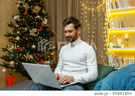 Successful businessman working at home for Christmas, man typing on laptop, freelancer working remotely, sitting on sofa near Christmas tree celebrating New Year holidays. Successful businessman working at home for Christmas, man typing on laptop, freelancer working remotely, sitting on sofa near Christmas tree celebrating New Year holidays. 95601804
