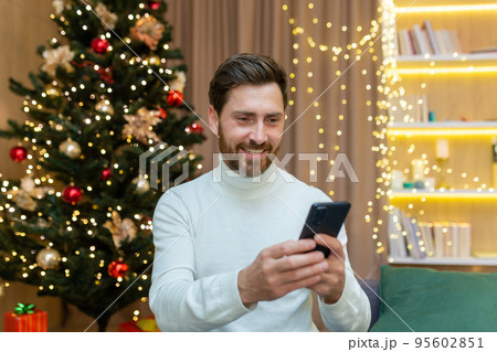 Cheerful man at home uses the phone, celebrates Christmas and New Year sitting on the sofa near the Christmas tree, types messages and browses online on the Internet. Cheerful man at home uses the phone, celebrates Christmas and New Year sitting on the sofa near the Christmas tree, types messages and browses online on the Internet. 95602851