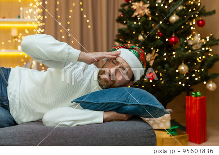 A man is tired for Christmas, in a New Year's hat, he is sleeping by the Christmas tree on the sofa at home in the living room after celebrating Christmas, he has a bad headache from a hangover. A man is tired for Christmas, in a New Year's hat, he is sleeping by the Christmas tree on the sofa at home in the living room after celebrating Christmas, he has a bad headache from a hangover. 95603836