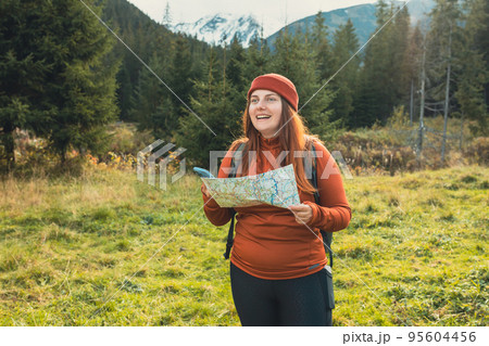 Stylish woman holding paper map and smartphone, wearing green backpack and red hat looking at mountain view while relaxing in nature. Travel and wanderlust concept. Amazing chill moment Stylish woman holding paper map and smartphone, wearing green backpack and red hat looking at mountain view while relaxing in nature. Travel and wanderlust concept. Amazing chill moment 95604456