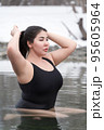 Large size young adult model with big breast in black one-piece bathing swimsuit sitting in outdoors pool at balneotherapy spa, hot springs resort. Woman raised hands straightens hair, looking away 95605964