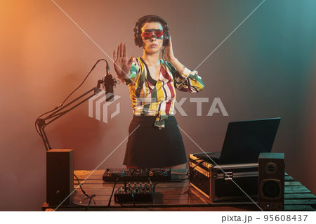 Young artist doing stop sign with palm and listening to sounds in headphones, mixing music at turntables. Cool female DJ showing rejection and denial gesture, forbidden or refusal. 95608437