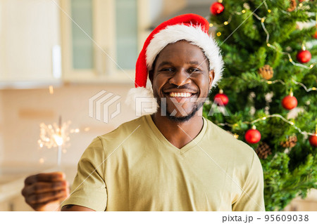 african american man in red Santa hat holding sparkler near christmas tree at home african american man in red Santa hat holding sparkler near christmas tree at home 95609038