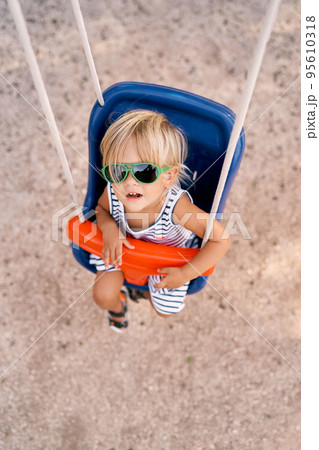 Little girl in sunglasses sits on a swing and looks up with her mouth open. High quality photo Little girl in sunglasses sits on a swing and looks up with her mouth open. High quality photo 95610318