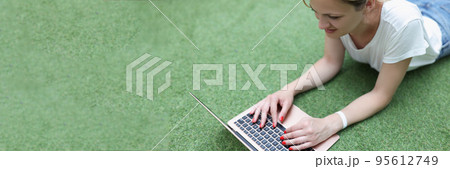 Smiling blonde woman working on laptop lay on grass, remote job, female freelancer outdoors 95612749