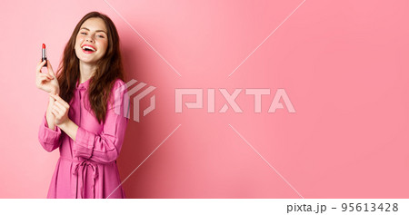 Beautiful woman smiling, applying lipstick and make up for party, standing against pink background, getting ready for going out 95613428