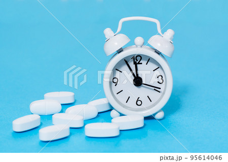 White alarm clock and white oval pills next to it on a blue background. Taking medicine at the right time. 95614046