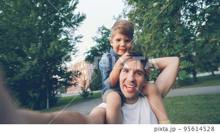 POV of Happy father carrying his smiling son on neck and making video selfie at smartphone camera during walk in park 95614528