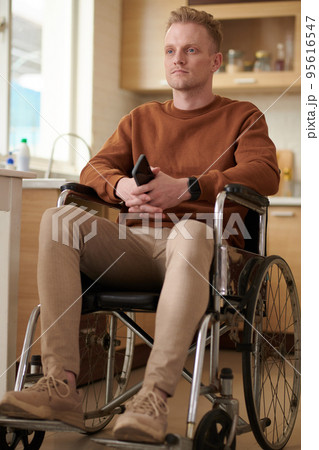 Portrait of serious young man with smartphone sitting in wheelchair and looking away 95616547