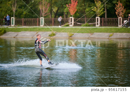 Wakeboarder surfing on lake. Young man surfer having fun wakeboarding in the cable park. Water sport, outdoor activity concept. Wakeboarder surfing on lake. Young man surfer having fun wakeboarding in the cable park. Water sport, outdoor activity concept. 95617155