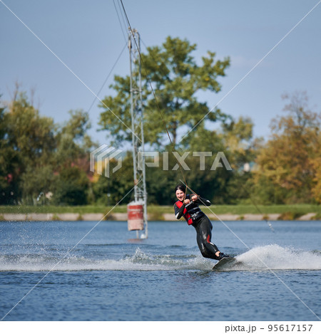 Wakeboarder surfing on lake. Young man surfer having fun wakeboarding in the cable park. Water sport, outdoor activity concept. Wakeboarder surfing on lake. Young man surfer having fun wakeboarding in the cable park. Water sport, outdoor activity concept. 95617157