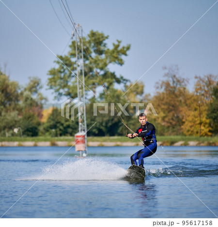 Wakeboarder surfing on lake. Young man surfer having fun wakeboarding in the cable park. Water sport, outdoor activity concept. Wakeboarder surfing on lake. Young man surfer having fun wakeboarding in the cable park. Water sport, outdoor activity concept. 95617158