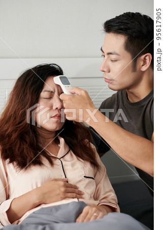 Caring husband helping sick wife to measure body temperature with electronic thermometer 95617905