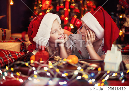 Little girl and boy in pajamas and Santa's hats lying near Christmas tree and looking to each other  95618459