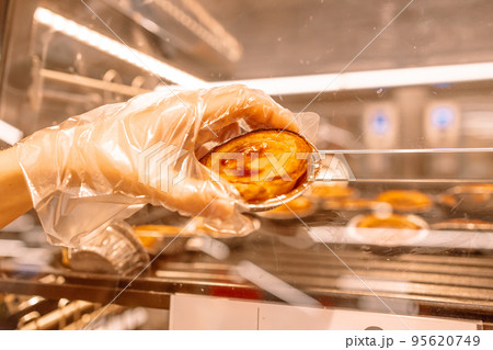 Traditional portuguese dessert. Woman's hand in a disposable glove takes Pastel de nata. Bakery shop window display. Traditional portuguese dessert. Woman's hand in a disposable glove takes Pastel de nata. Bakery shop window display. 95620749