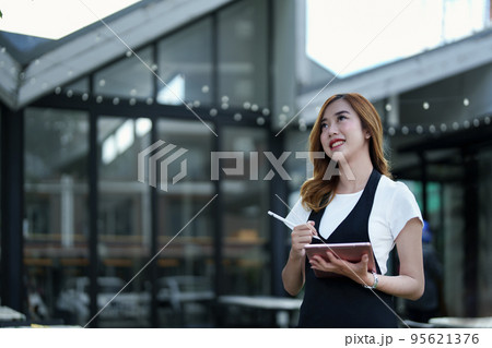 Starting and opening a small business, a young Asian woman showing a smiling face holding a tablet in an apron standing in front of a coffee shop bar counter. Business Owner, Restaurant, Cafe concept. 95621376