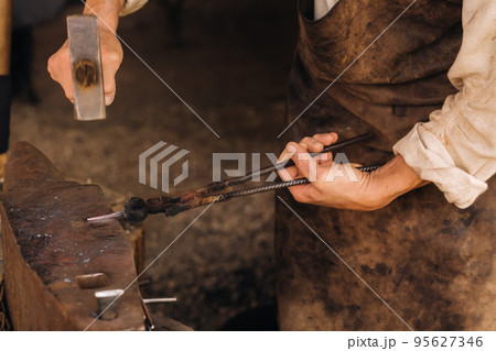 A blacksmith manually forges red-hot metal on an anvil with a hammer A blacksmith manually forges red-hot metal on an anvil with a hammer 95627346