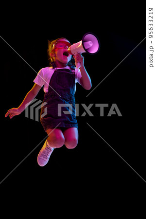 Thrilled elementary school age girl, pupil jumping with megaphone isolated over dark background. Concept of children emotions, fashion, facial expressions Thrilled elementary school age girl, pupil jumping with megaphone isolated over dark background. Concept of children emotions, fashion, facial expressions 95633219
