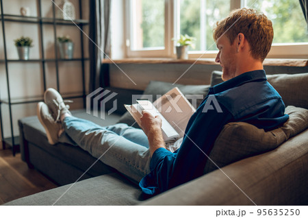 A ginger man sitting at the window and reading a book 95635250