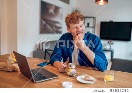 Ginger young man sitting in th kitchen and making sandwich with chocolate paste Ginger young man sitting in th kitchen and making sandwich with chocolate paste 95635256