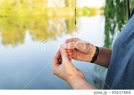 The concept of bait for fish. Close-up of a fisherman's male hand stringing bait on a fishing rod against a blue lake. 95637868