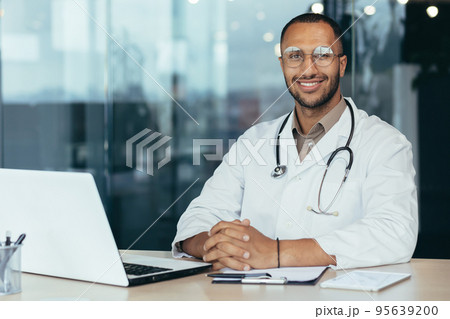Portrait of young hispanic male pediatrician doctor. Sitting in the office at a table with documents and a laptop, wearing glasses and a stethoscope in a white coat, looking at the camera, smiling. 95639200