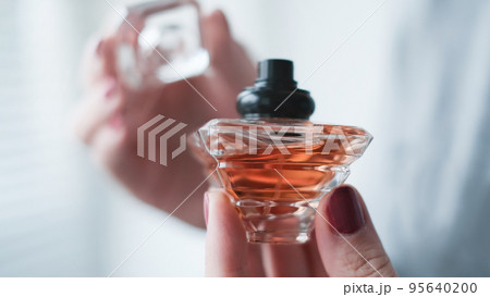 Perfume bottle in a female hand against the background of light room interior. 95640200