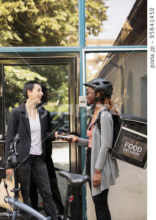 Smiling client paying for office food delivery with credit card, pos contactless payment. African american courier with backpack holding terminal, giving restaurant meal to customer 95641450