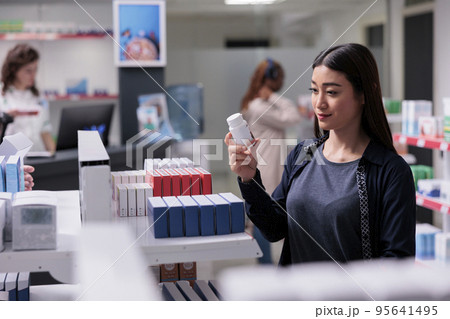 Woman client holding drugs bottle looking at pharmaceutical prescription, buying medication treatment for immune system in pharmacy. Drugstore shelves full with vitamin, supplements ready for purchase 95641495