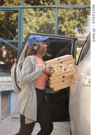 Pizzeria courier delivering pizza to office by car, side view. Lunch delivery service employee in headphones holding fastfood boxes stack, standing near company building outdoors 95641496
