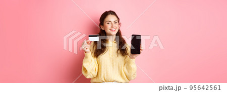 Technology and online shopping. Portrait of beautiful lady showing mobile phone screen and plastic credit card, smiling pleased, recommend app, standing over pink background 95642561