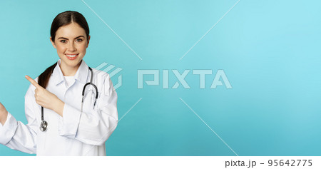 Portrait of smiling medical worker, girl doctor in white coat with stethoscope, pointing fingers left, showing medical clinic advertisement, torquoise background Portrait of smiling medical worker, girl doctor in white coat with stethoscope, pointing fingers left, showing medical clinic advertisement, torquoise background 95642775