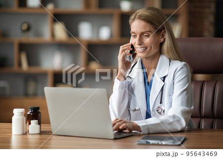 Smiling Doctor Woman Talking On Cellphone And Using Laptop At Workplace 95643469