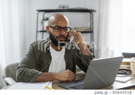 Latin male freelancer calling by cellphone while working online on laptop, having phone conversation at home 95643518