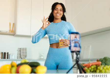 Young sporty black lady filming video about healthy eating on smartphone, holding glass of juice and gesturing okey Young sporty black lady filming video about healthy eating on smartphone, holding glass of juice and gesturing okey 95643555