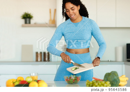 Slender african american woman in sportswear standing in the kitchen and preparing fresh vegetable salad Slender african american woman in sportswear standing in the kitchen and preparing fresh vegetable salad 95643586