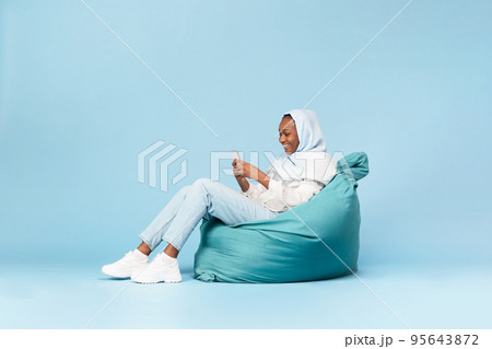 Positive black muslim woman chilling in beanbag chair with smartphone, chatting with friends in social networks Positive black muslim woman chilling in beanbag chair with smartphone, chatting with friends in social networks 95643872