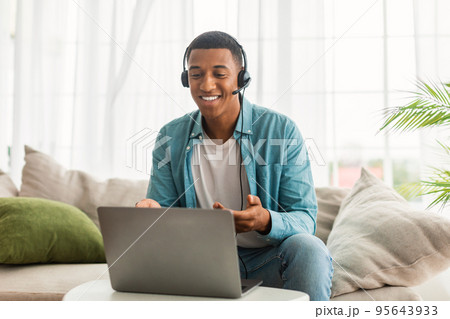 Cheerful young black man in jeans with headphones has video call on laptop, supports client, chats remotely 95643933