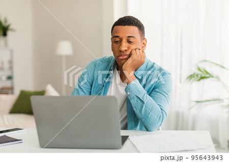 Sad tired bored young black guy in casual looks at laptop in light living room office interior, empty space 95643943