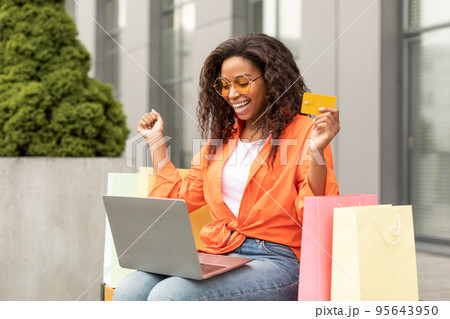 Satisfied millennial african american female in glasses with many bags with purchases, rejoice to win, shows credit card 95643950