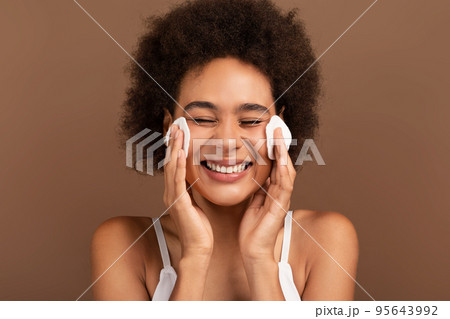 Happy young mixed race lady with curly hair removes makeup from face with cotton pad Happy young mixed race lady with curly hair removes makeup from face with cotton pad 95643992