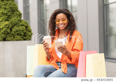Happy millennial african american lady in casual, glasses with many bags enjoy cup of drink takeaway, typing on phone Happy millennial african american lady in casual, glasses with many bags enjoy cup of drink takeaway, typing on phone 95643995