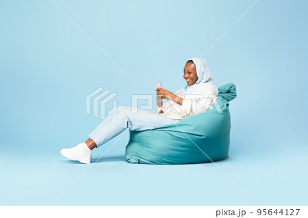 Excited black woman in hijab sitting on beanbag chair and using modern cellphone, using entertaining mobile application Excited black woman in hijab sitting on beanbag chair and using modern cellphone, using entertaining mobile application 95644127