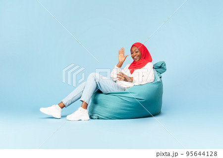 Full length of happy black muslim woman sitting in beanbag chair with smartphone and having video chat with friend 95644128