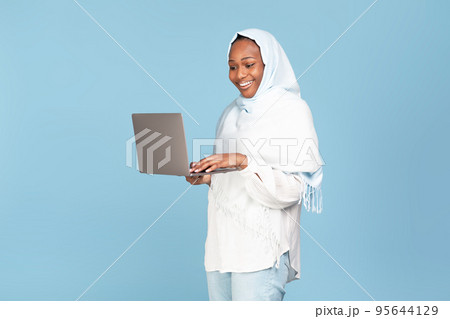 Freelance and online education for muslim women concept. Happy black woman in hijab holding and typing on laptop 95644129