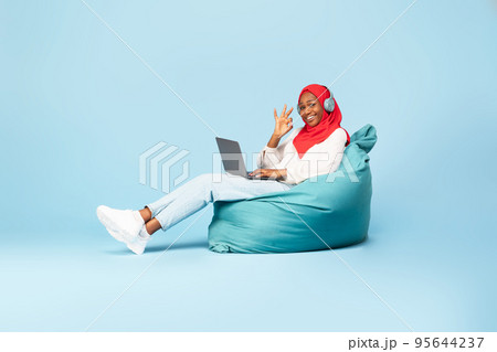 Black muslim woman sitting in beanbag chair with laptop and showing okay gesture, recommending online work or education Black muslim woman sitting in beanbag chair with laptop and showing okay gesture, recommending online work or education 95644237