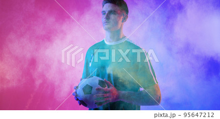 Caucasian male player holding soccer ball looking away standing against smoky background, copy space 95647212
