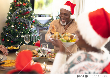 Happy african american family wearing santa hats, sitting at table and eating dinner 95648950